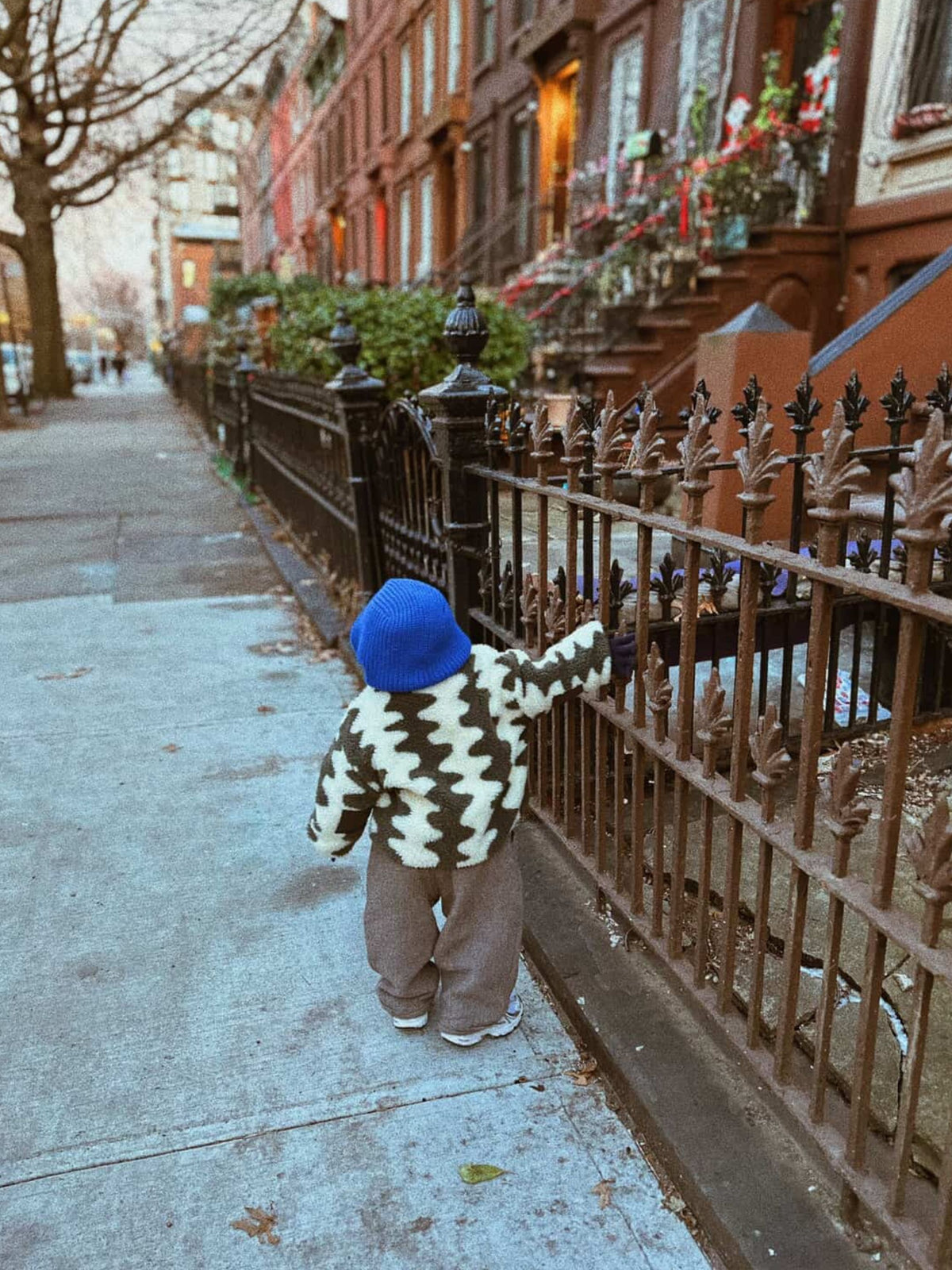 Charcoal | A child in a blue hat stands outdoors on a leafy city sidewalk, facing away from the camera, cozy in the WAVELENGTH SHERPA JACKET featuring a black and white pattern and quilted interior.