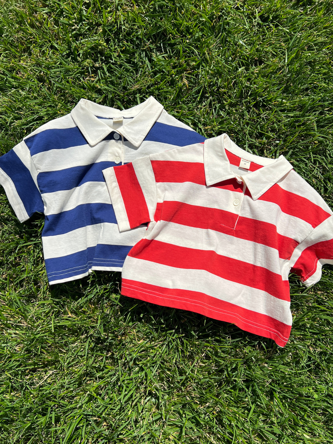 Two RUGBY COLLAR TEE shirts—one blue and white, the other red and white—both short-sleeved with horizontal stripes, 100% cotton from a Korean kids brand, are lying on green grass.