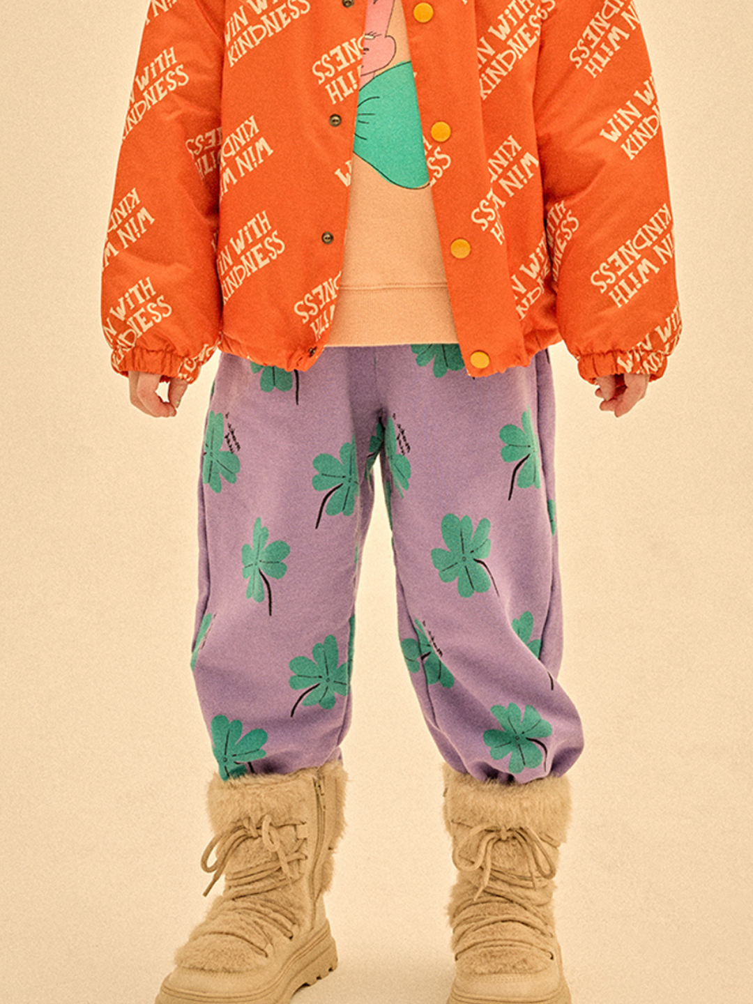 A child dressed in orange jacket with white "WITH KINDNESS" text, peach shirt, LUCKY CLOVER ALADDIN PANTS featuring green clovers, and beige furry boots stays