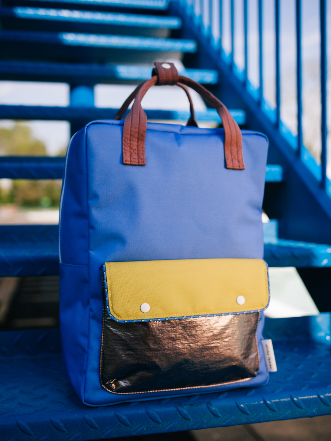 The LARGE BETTER TOGETHER ENVELOPE BACKPACK, in blue with brown handles and a yellow and metallic bronze front pocket, sits on a blue metal staircase and features adjustable shoulder straps for comfort.