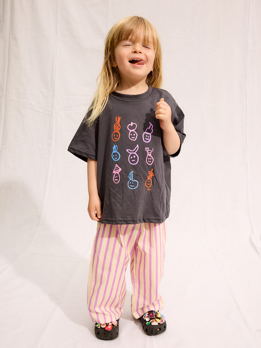 A child is wearing a Charcoal Fruit face tee with Pine Pants and black crocs