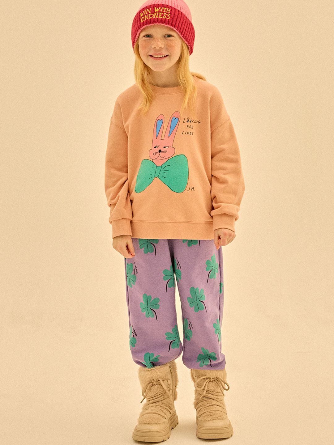 A young girl in a pink beanie, peach bunny sweatshirt, LUCKY CLOVER ALADDIN PANTS, and beige boots beams at the camera—her bright outfit shows style