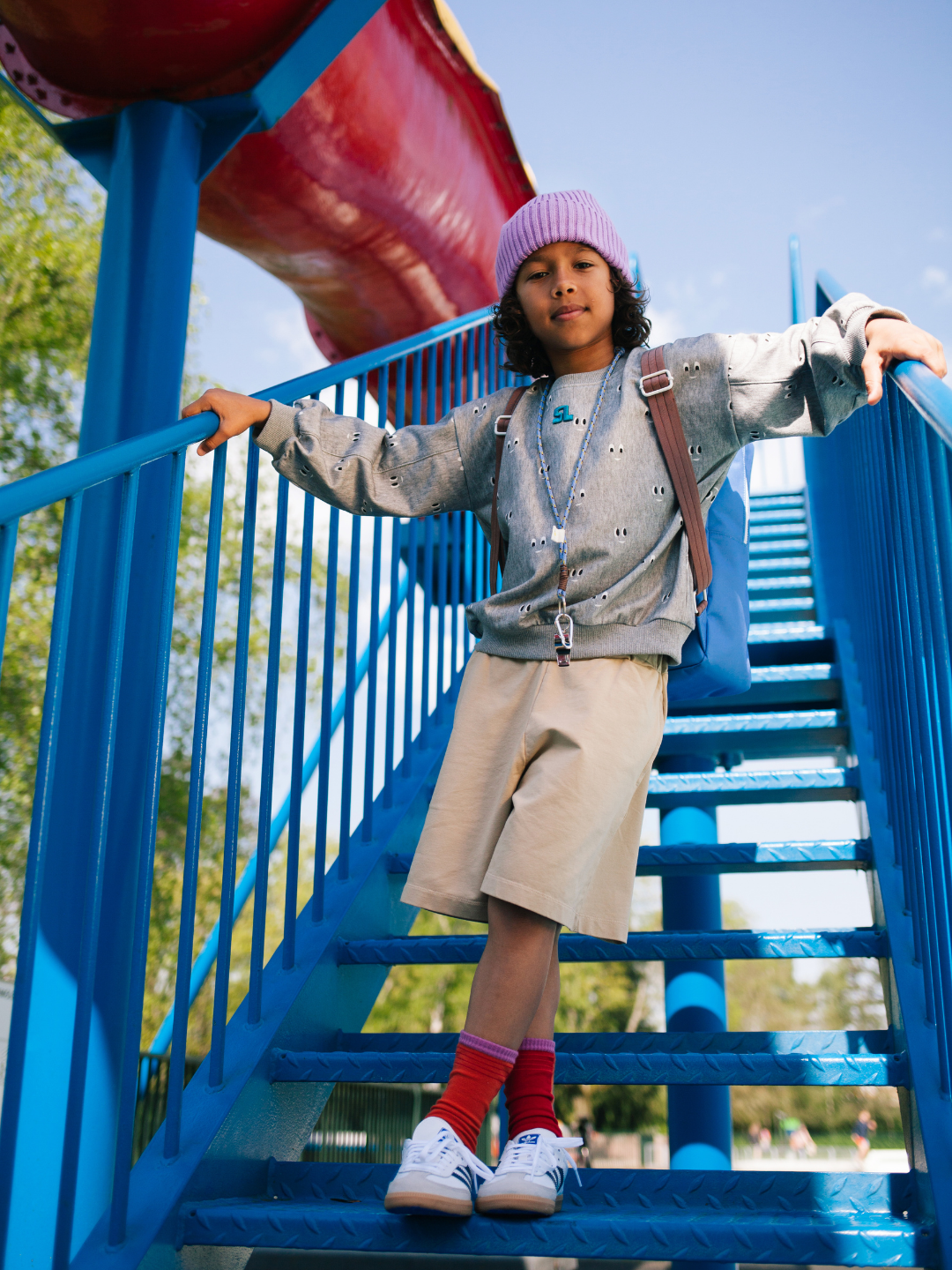 A child in a grey sweatshirt, beige shorts, red socks, and a purple beanie stands on blue metal stairs outdoors, holding the railing and carrying the LARGE BETTER TOGETHER ENVELOPE BACKPACK with adjustable shoulder handles.