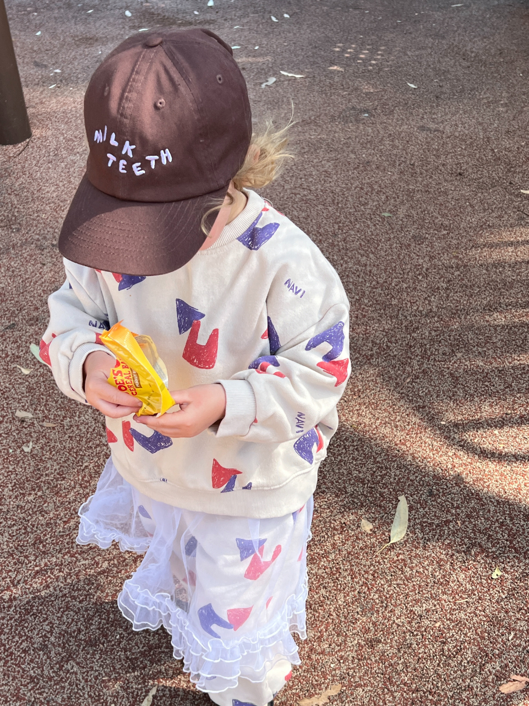 Brown | Child wearing the Milk Teeth baseball cap in brown with pale purple Milk Teeth lettering, outdoors at a park with a brown floor. They are wearing a cream sweatshirt printed with purple and red shapes, a white tutu skirt, and eating a snack bar in a yellow wrapper.