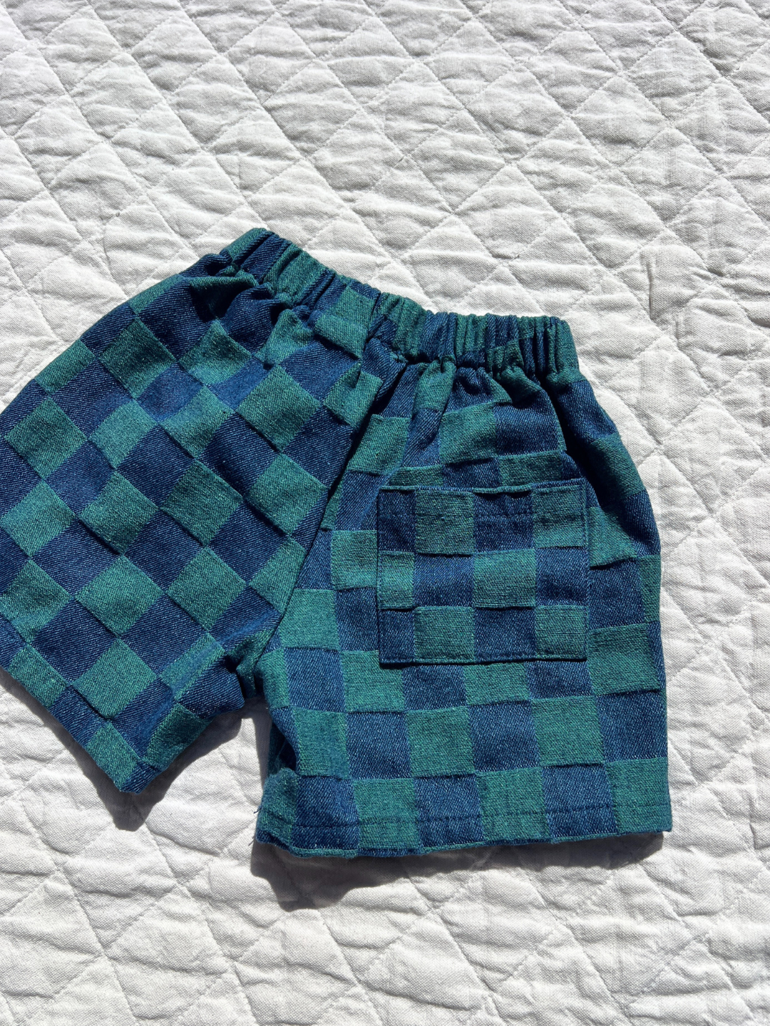 Green | TWO TONE DENIM CHECK SHORTS: Blue and green checkerboard jacquard shorts with an elastic waistband and back pocket, from a Korean kids brand, shown on a white quilted surface.