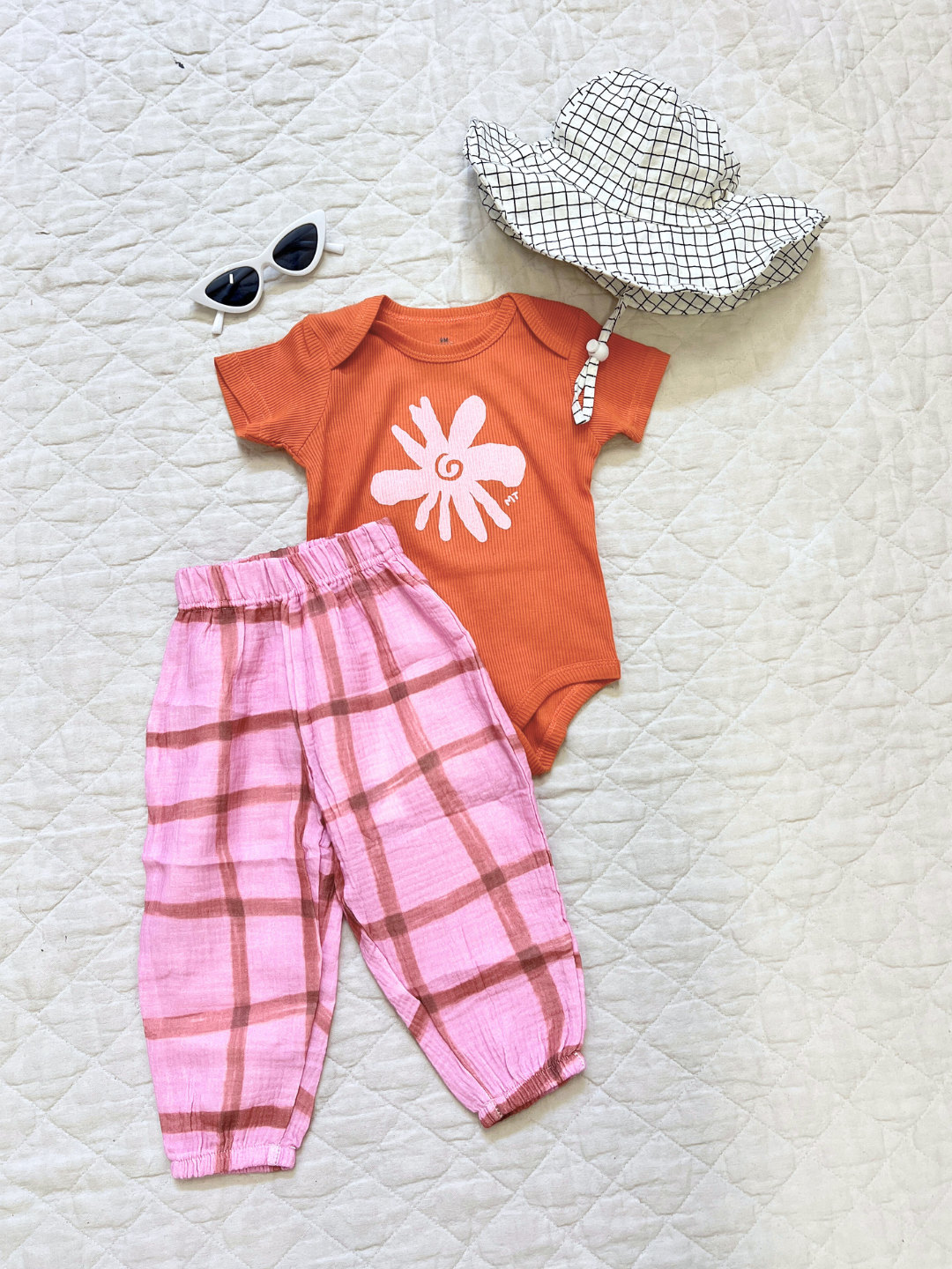 White | A flat outfit with the baby off the grid hat in white, paired with pink check pants and a flower onesie, laid on a white cloth.