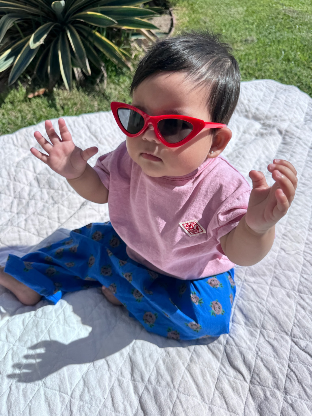A baby wears the Pascal pink tee with blue floral pants and red sunglasses, seated on a white blanket on green grass.