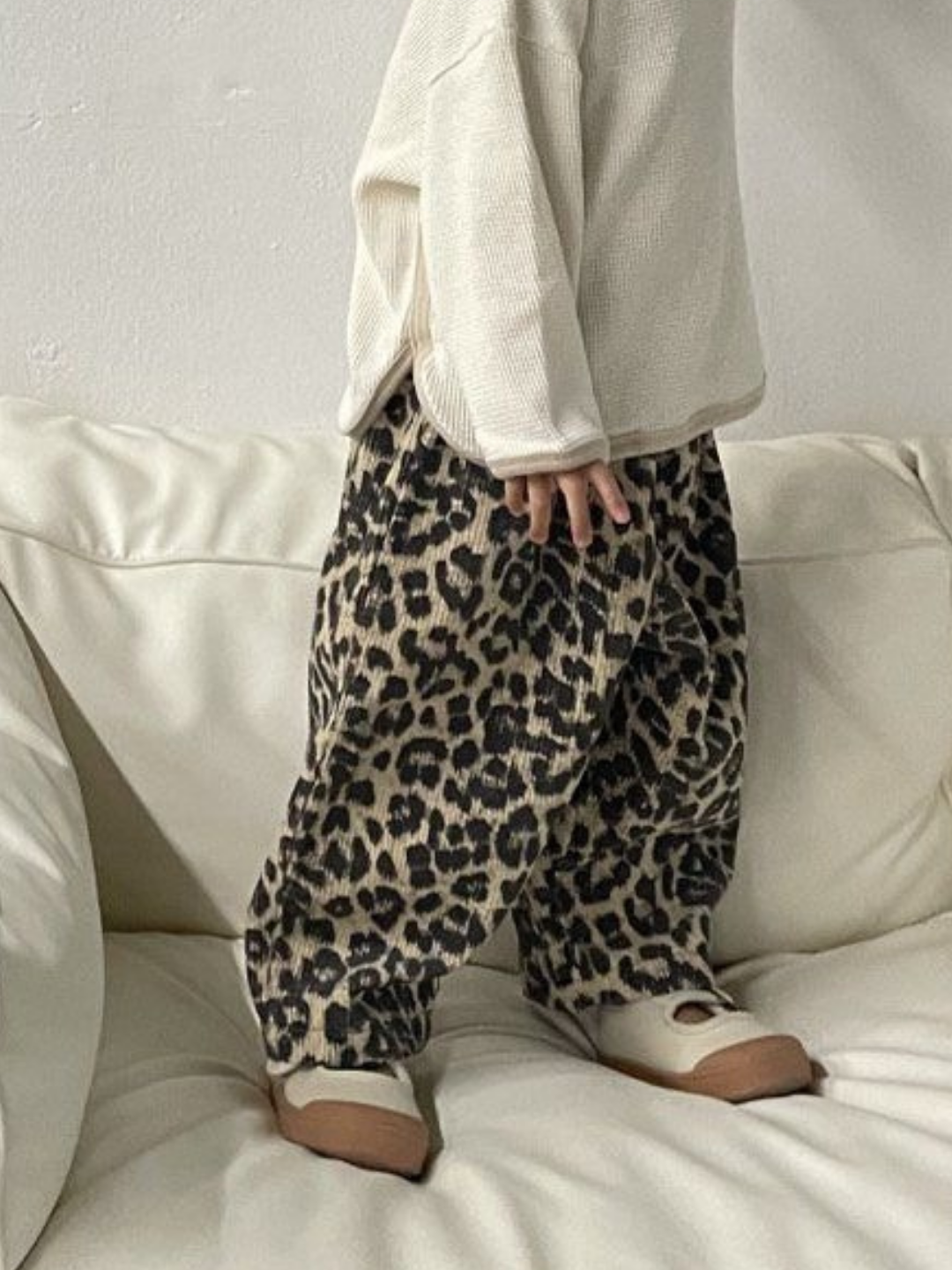Light | A child stands on a cream couch wearing a beige long-sleeve top, LEOPARD CORDUROY PANTS, and beige shoes from a popular Korean brand.