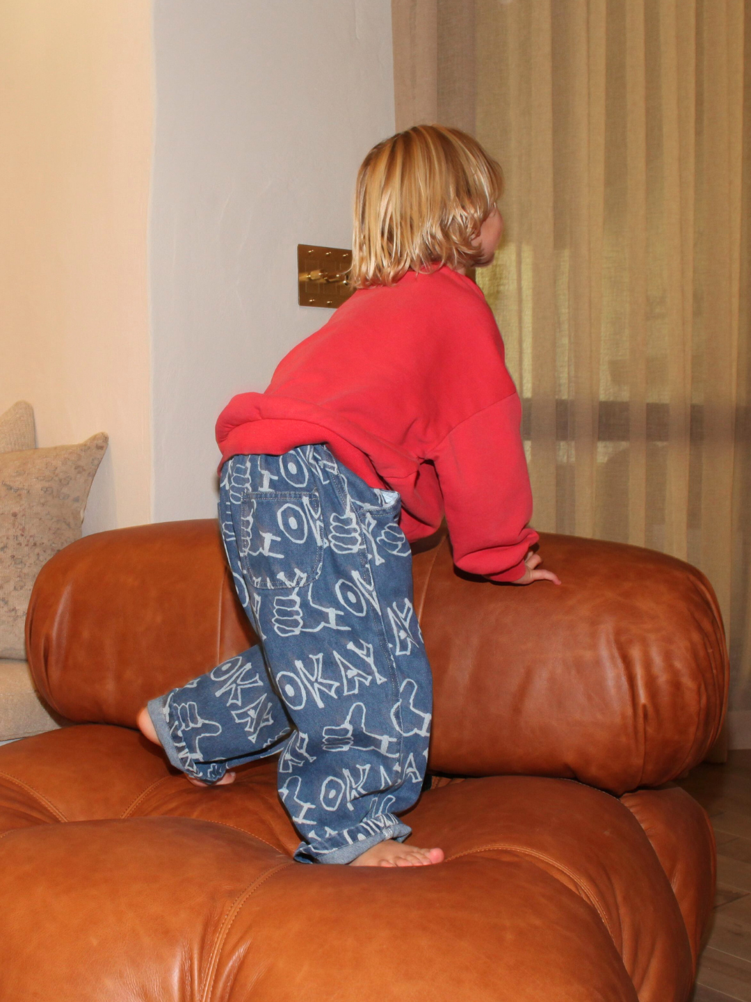 Blue | A young blonde-haired child in a red sweatshirt and OKAY JEANS stands barefoot on a brown leather couch, gazing at a window with beige curtains.