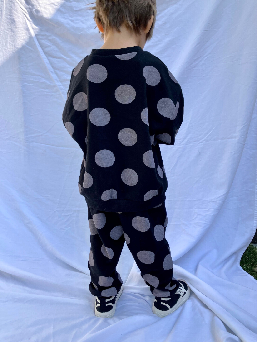 Back view of child facing away from camera, wearing a sweat set in black with large grey polka dots. The set consists of a crewneck sweatshirt and a matching sweatpant, with an oversized fit. He wears black velcro sneakers with white trim and is standing on a white sheet backdrop.