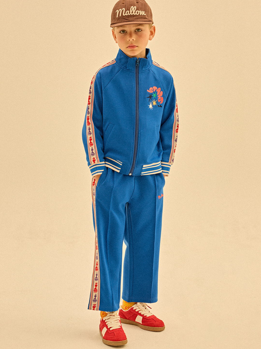 A young boy in a CHESS TRACK JACKET with striped side panels, a brown cap, and red sneakers stands against a beige background