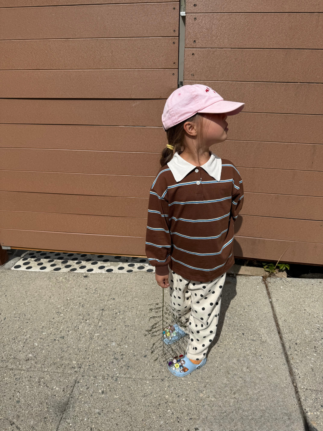 Child wearing a striped shirt and pink cap standing against a brown wall.