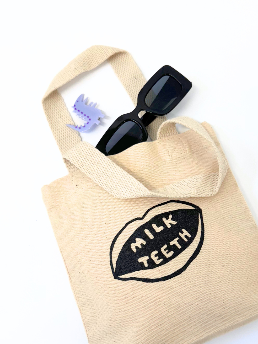 Milk Teeth mini tote laid flat with sunglasses and Dino clip