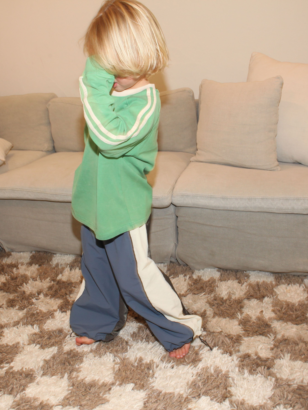 A blonde-haired child stands on a patterned rug in front of a sofa, wearing an oversized green HALFTIME LONGSLEEVE and blue pants, with one hand partially covering their face.
