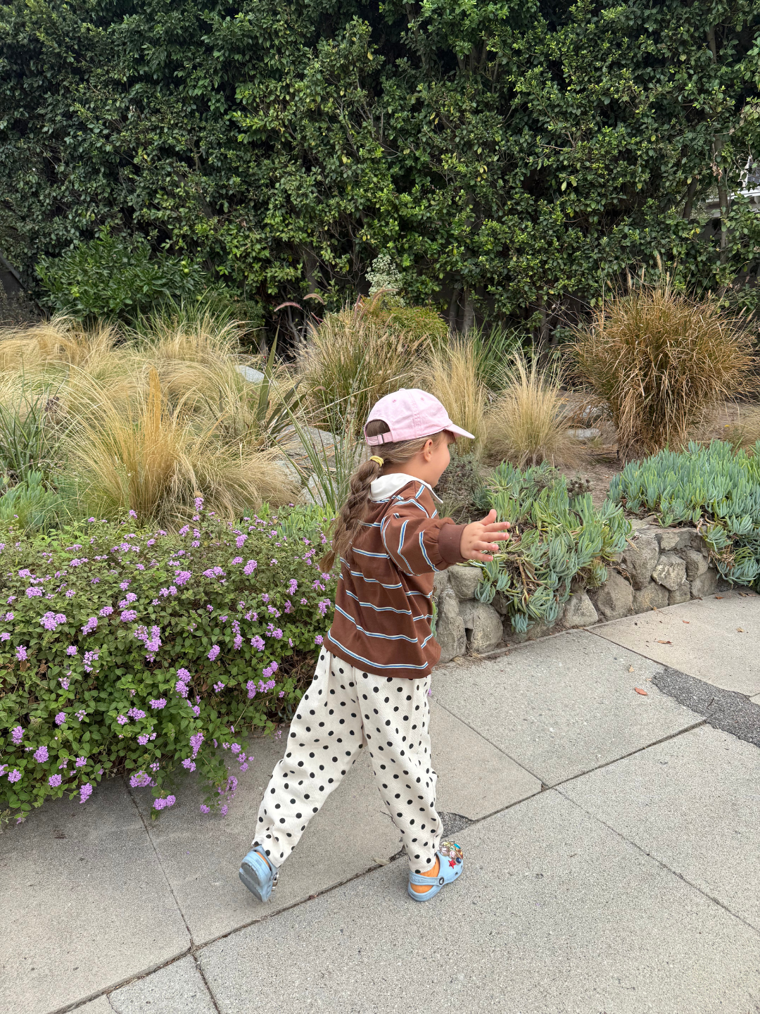 Child in a striped sweater and polka dot pants standing on a sidewalk with greenery in the background