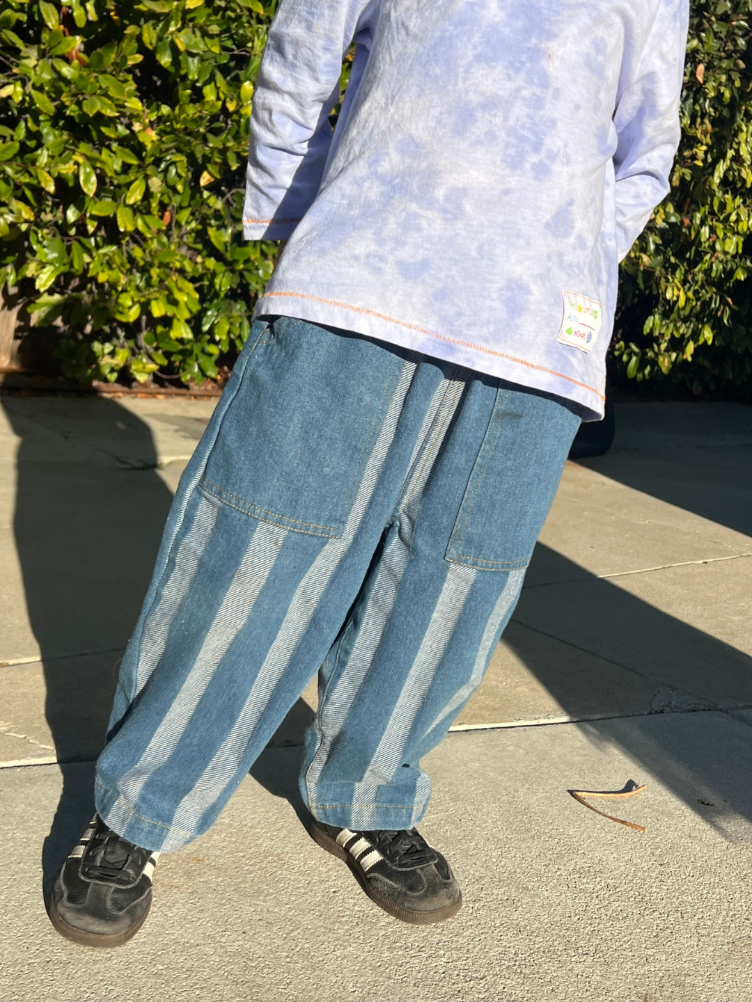 Medium Wash | A person wearing STRIPE DOUBLE TROUBLE JEANS, black sneakers, and a light purple tie-dye shirt stands on the pavement; a leafy bush provides the backdrop.