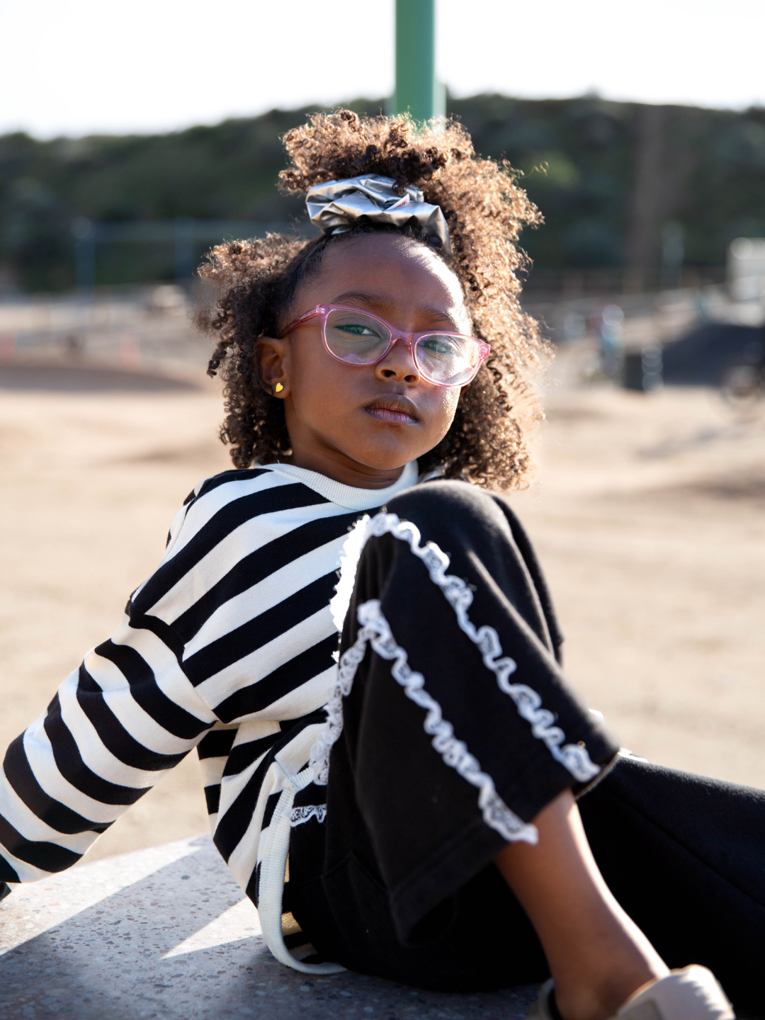 Girl wearing the stardust oversized scrunchie in metallic silver. She has a half ponytail in her curly hair and wears pink glasses, gold heart earrings and a black and white striped top and black track pants with white ruffles down the side. She is seated outdoors in a park with one leg up.