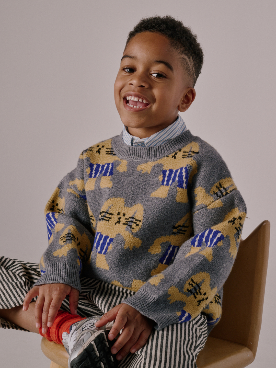 Gray | A young boy smiles while seated, wearing the MAX SWEATER featuring gray fabric with yellow and blue bunny patterns, along with a collared shirt, striped pants, and sneakers.