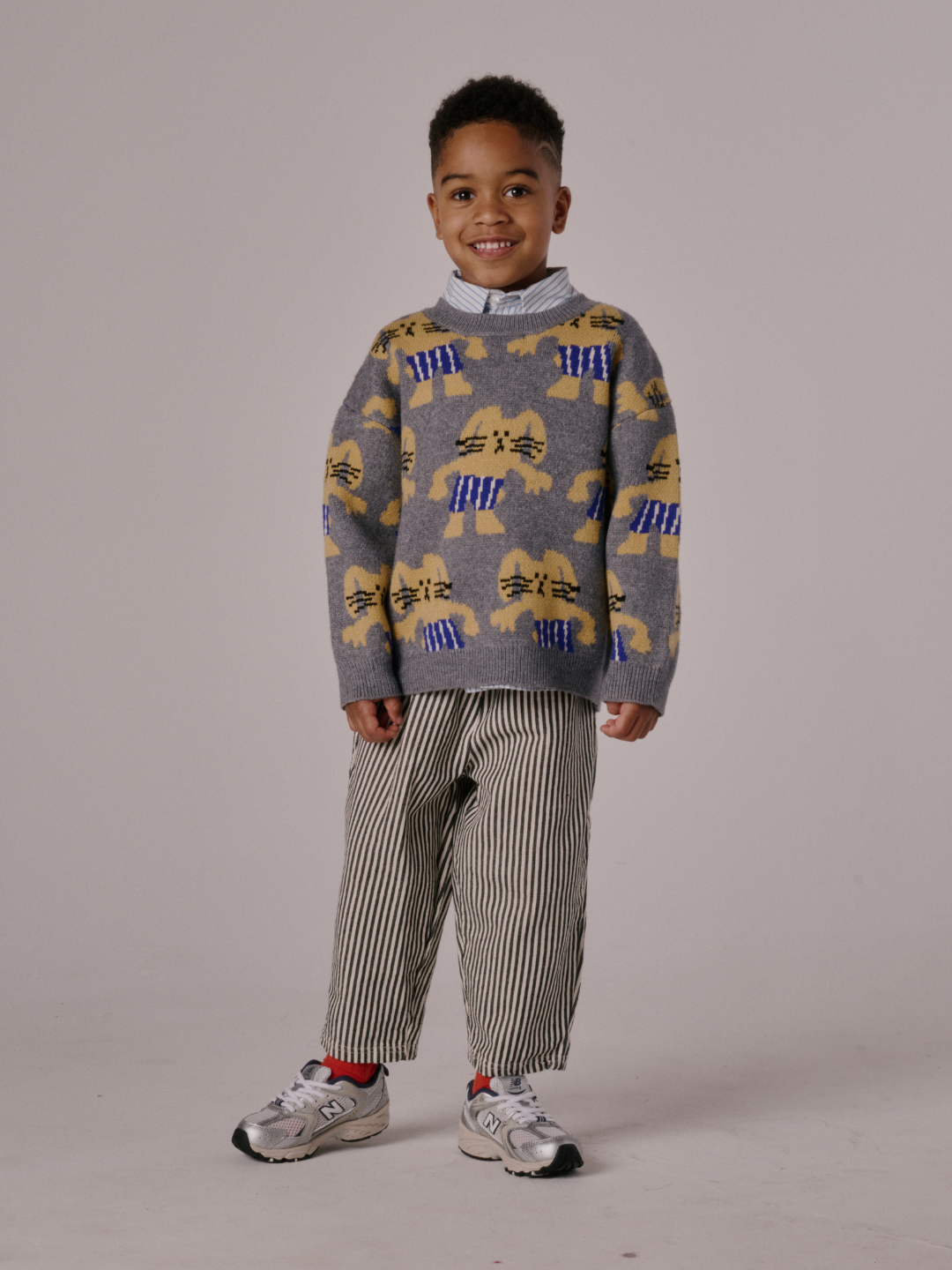 Gray | Young child standing and smiling, wearing the MAX SWEATER (gray with bunny designs), striped pants, red socks, and white sneakers against a plain background.