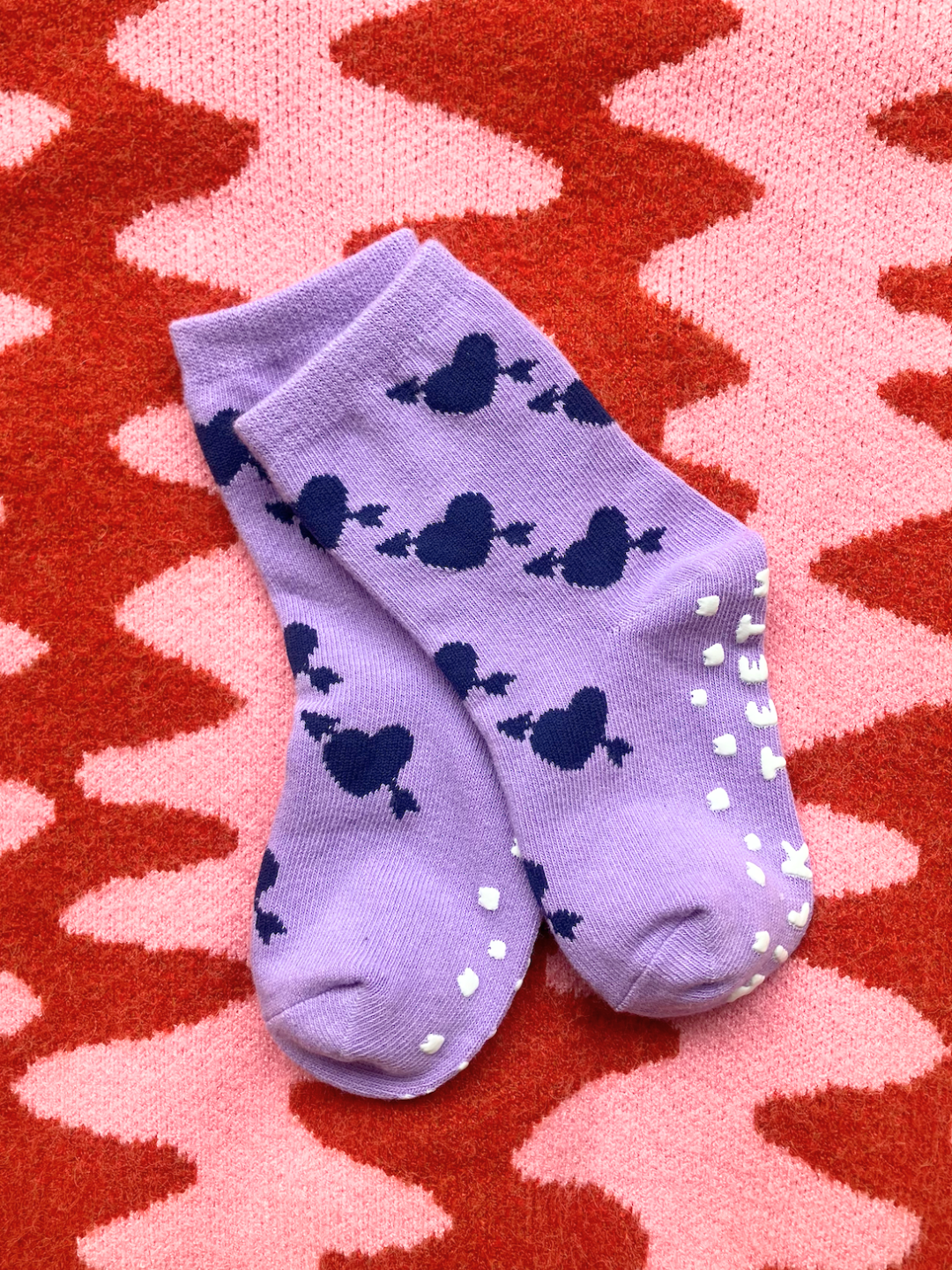 Violet Lovestruck | The GRIP SOCK, a plush pair of purple socks with navy blue heart patterns and white anti-skid dots on the soles, is displayed against a pink and red zigzag patterned background.