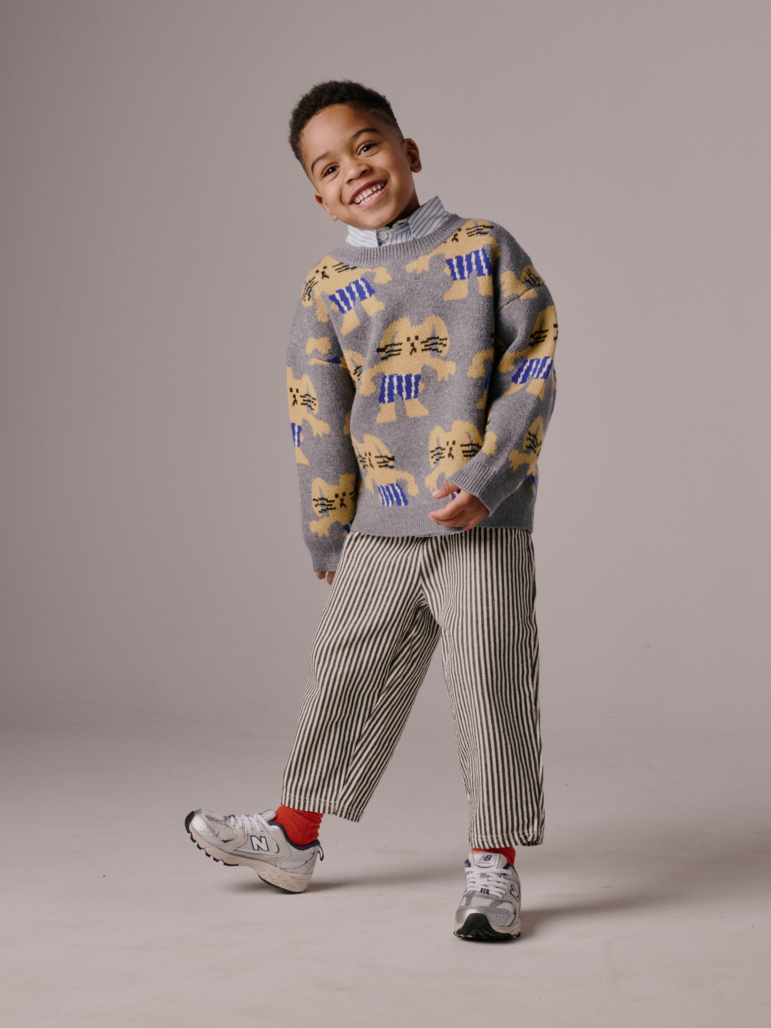 Gray | A smiling child stands indoors against a plain background, wearing the MAX SWEATER—a grey, bunny-patterned top—paired with striped pants, red socks, and grey sneakers.