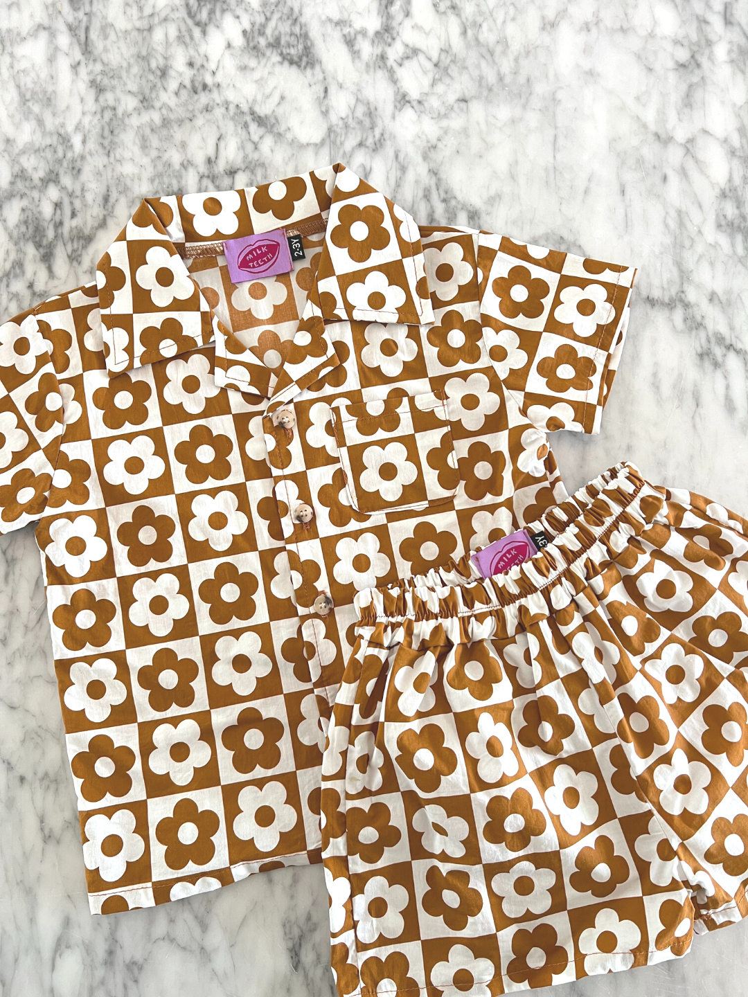Terracotta | Set of kids' shirt and shorts in a checkerboard pattern of terracotta brown and white flowers, laid on a table, front view