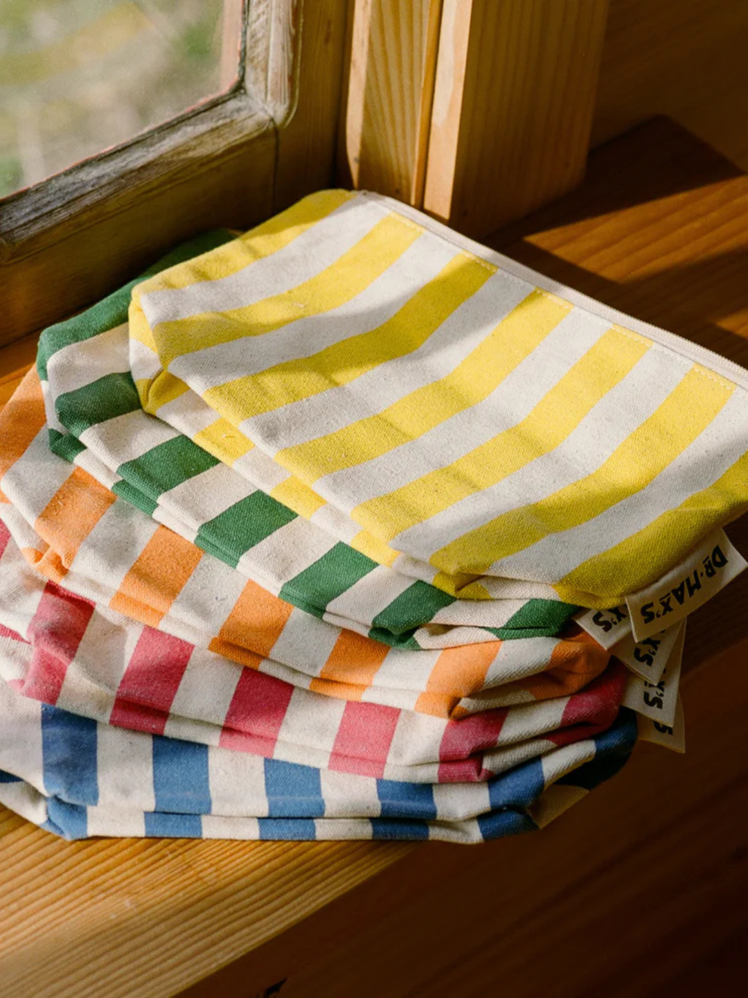 Four ORGANIC COTTON ZIPPER BAGs in yellow, green, red, and blue are stacked on a wooden surface by a window—ideal for organizing diaper bags and made from durable organic cotton.