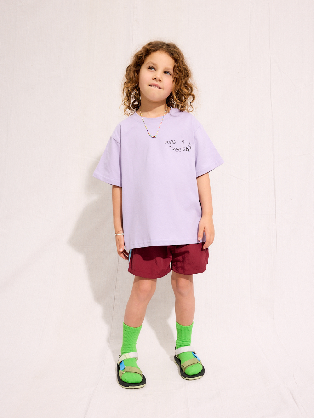 Purple | A child is wearing the purple Milk Teeth test with burgundy shorts and green socks and sandals