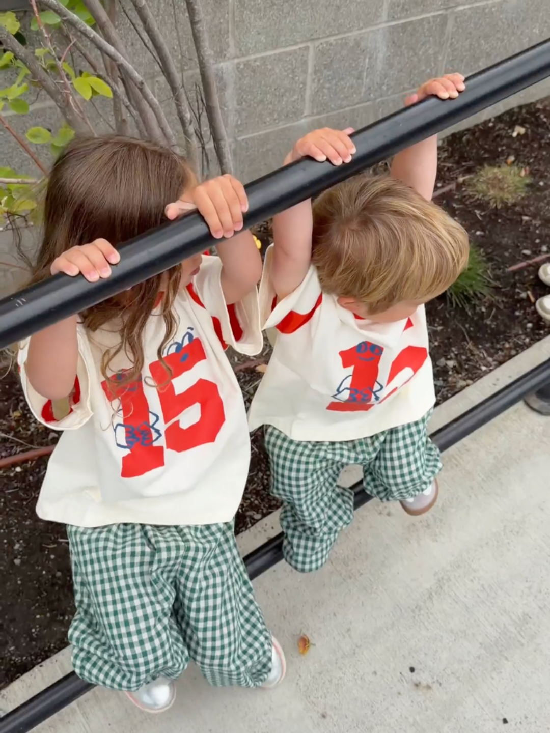 Two toddlers climbing on a black metal railing outdoors. Both are wearing matching outfits consisting of off-white t-shirts with a large red number '15' graphic, green and white gingham pants, and silver shoes.