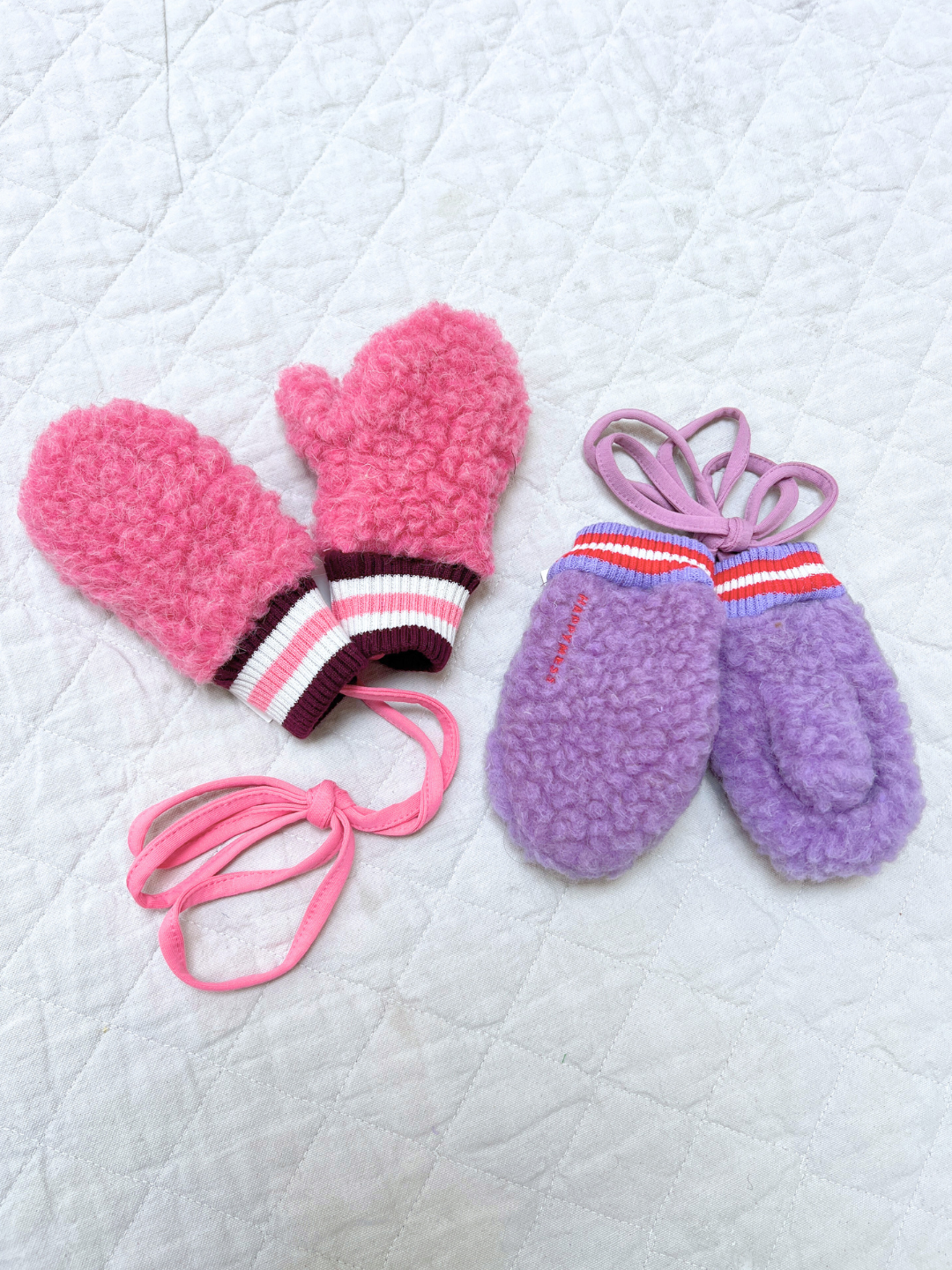 Lilac Cloud |Pair of pink and purple mittens with elastic bands on a white quilted background
