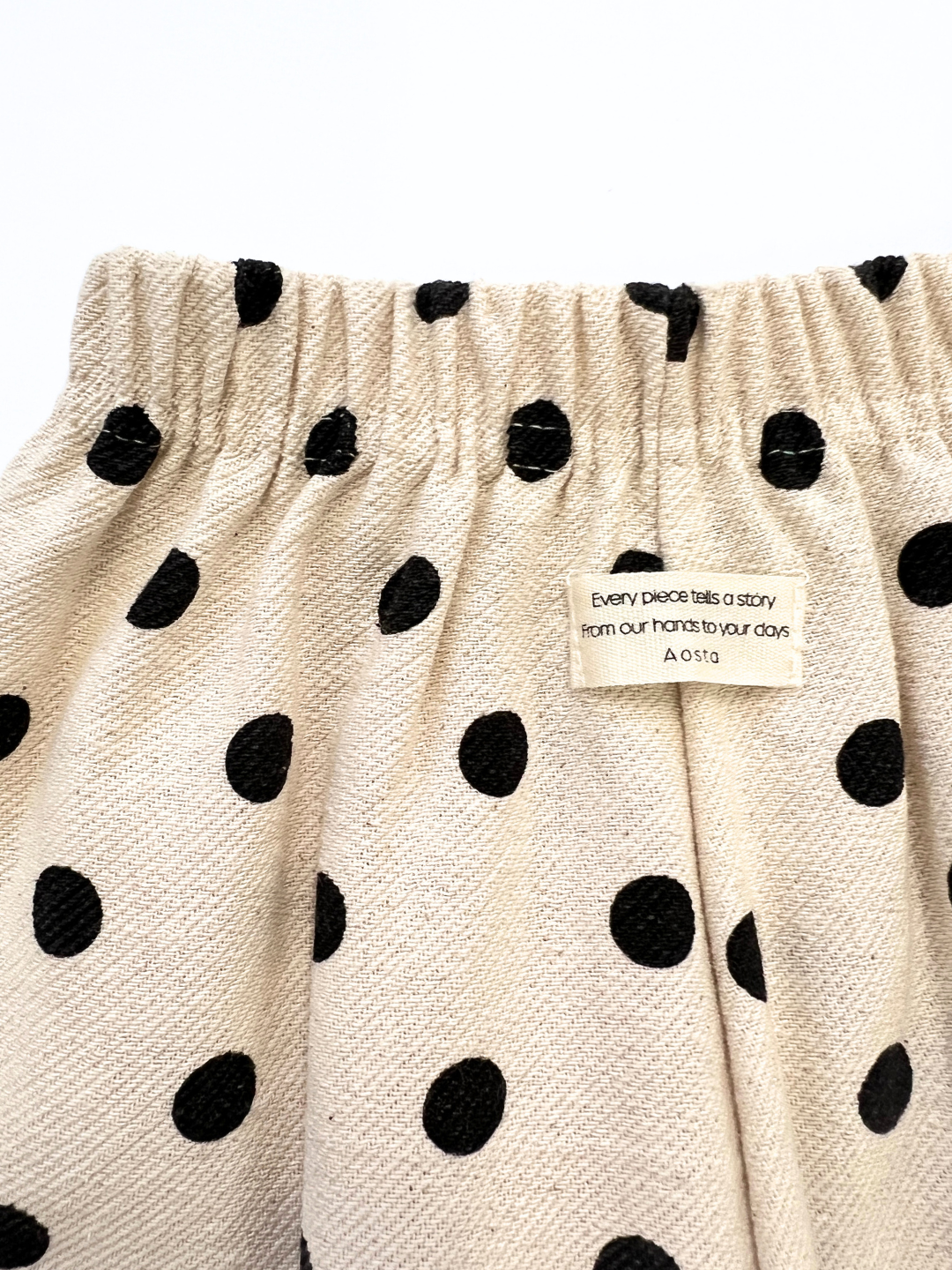 The POLKA DOT PANT features beige fabric with black polka dots, an elastic waistband, relaxed fit, double-layer cotton, and a label saying, "Every piece tells a story from our hands to your days. Aosta.