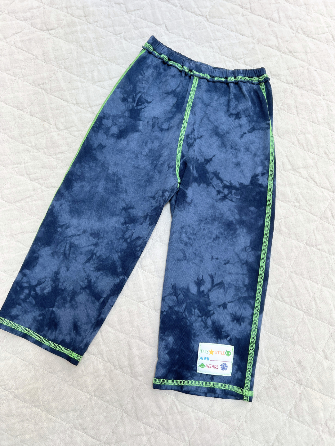 Pacific Blue | COSMO TIE DYE PANT for children in blue tie-dye with green stitching, shown flat on a quilted white surface. A patch near the hem says "THIS LITTLE KID WEARS." Crafted from soft cotton jersey for comfort and style.