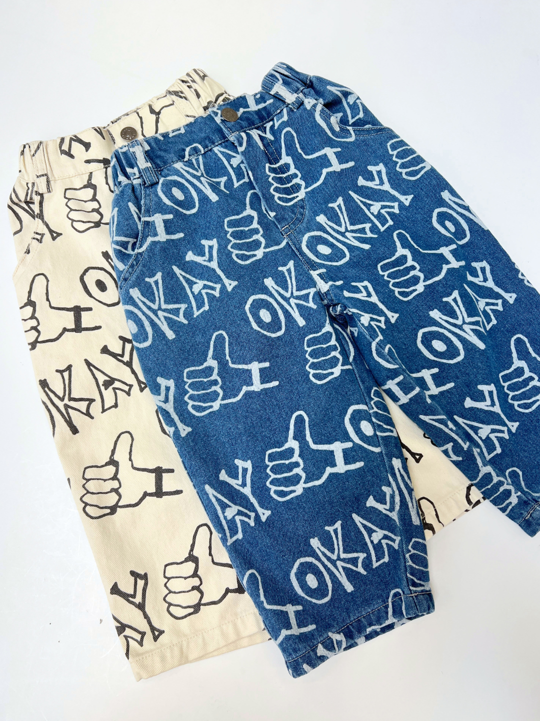 Two pairs of OKAY JEANS for kids—one white with black "OK" and thumbs-up prints, the other blue printed denim—are relaxed-fit jeans displayed side by side on a white background.