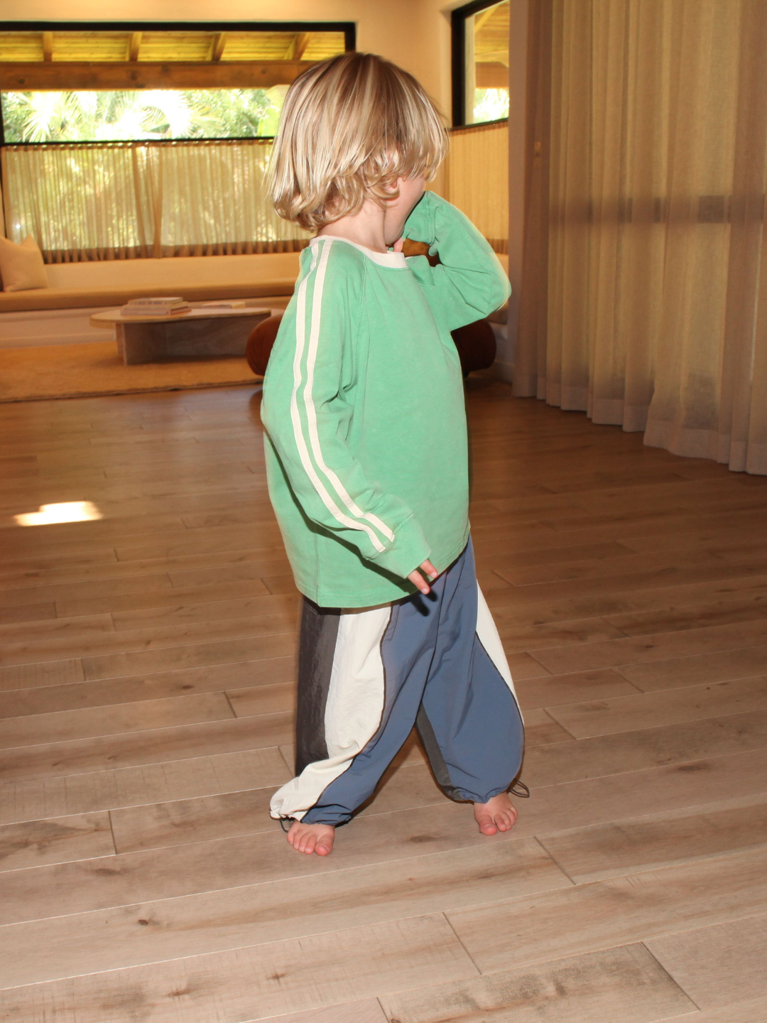 Green | A young child wears the HALFTIME LONGSLEEVE in green with an oversized fit and blue pants, standing barefoot on a wooden floor in a sunlit room, facing sideways with one hand to their face.