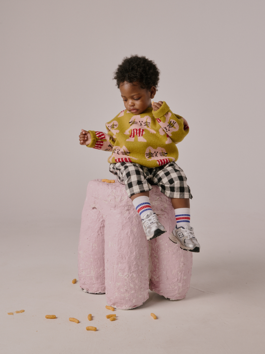 Chartreuse | A young child wears the MAX SWEATER while sitting on a pink, textured stool with snacks scattered around.