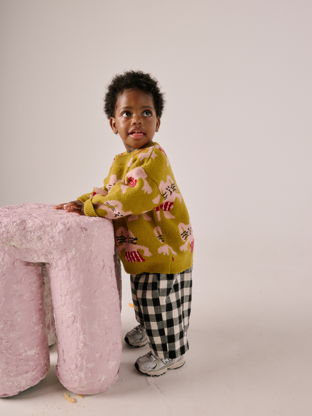 Chartreuse | A toddler wearing the yellow MAX SWEATER with bunny patterns, checkered pants, and sneakers stands beside a pink textured object in a studio setting.