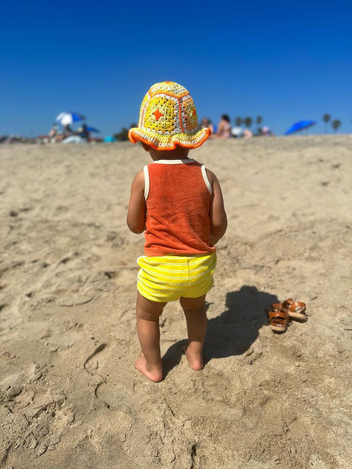 A young child wears the HAND-CROCHETED BUCKET HAT - 1-3Y, orange tank top, and yellow shorts while standing barefoot on a sandy South African beach. Nearby sandals, blue sky, and people are visible in the background.
