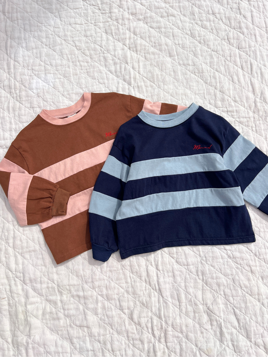 Three children's sweaters in brown, pink, and navy with light blue stripes on a textured white surface.