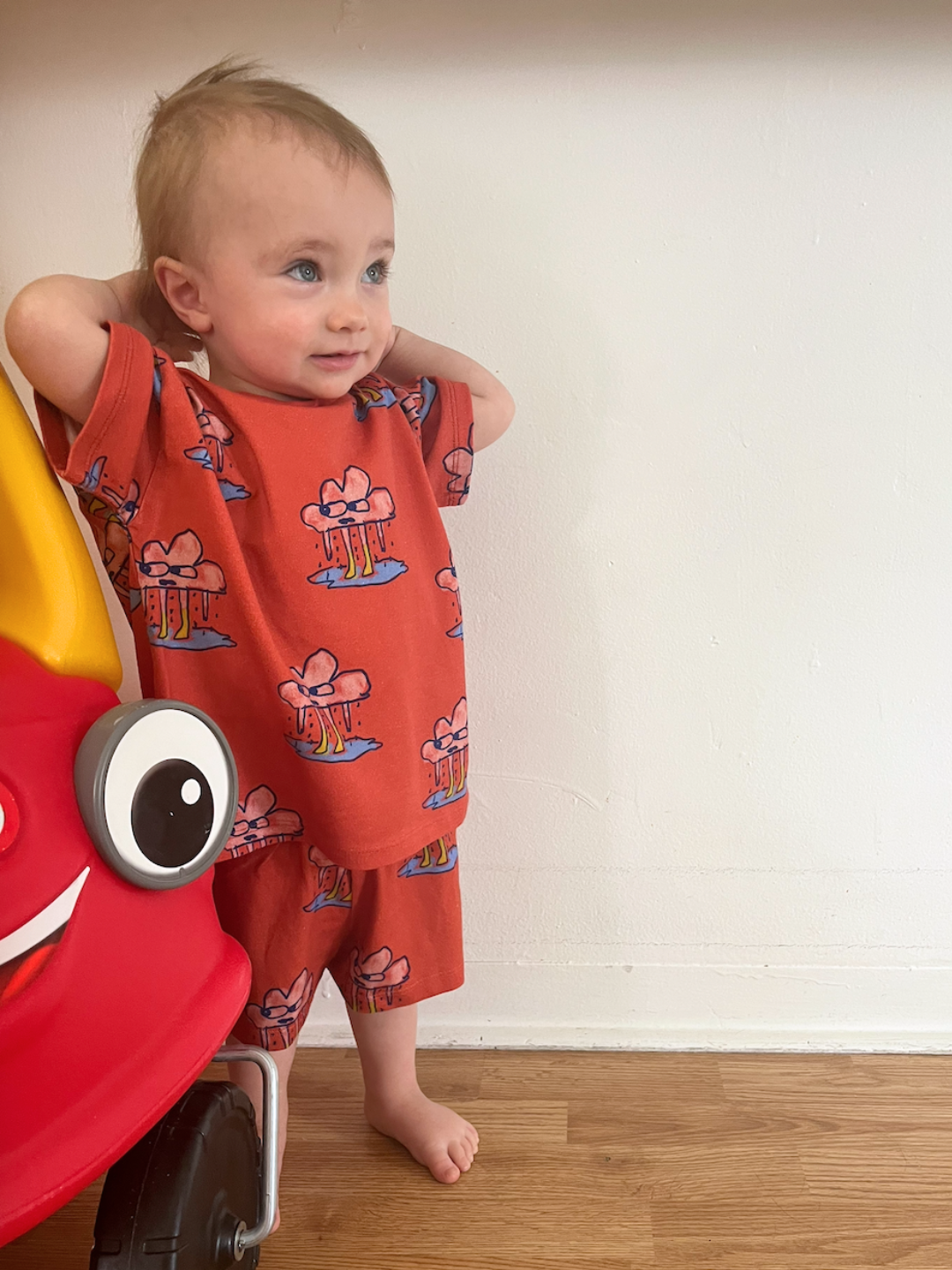 Red | A toddler indoors against a white wall, standing next to a red play car, wearing the RAINY DAY SHORT SET,a children’s unisex matching t-shirt and shorts set in red, in soft organic cotton.
