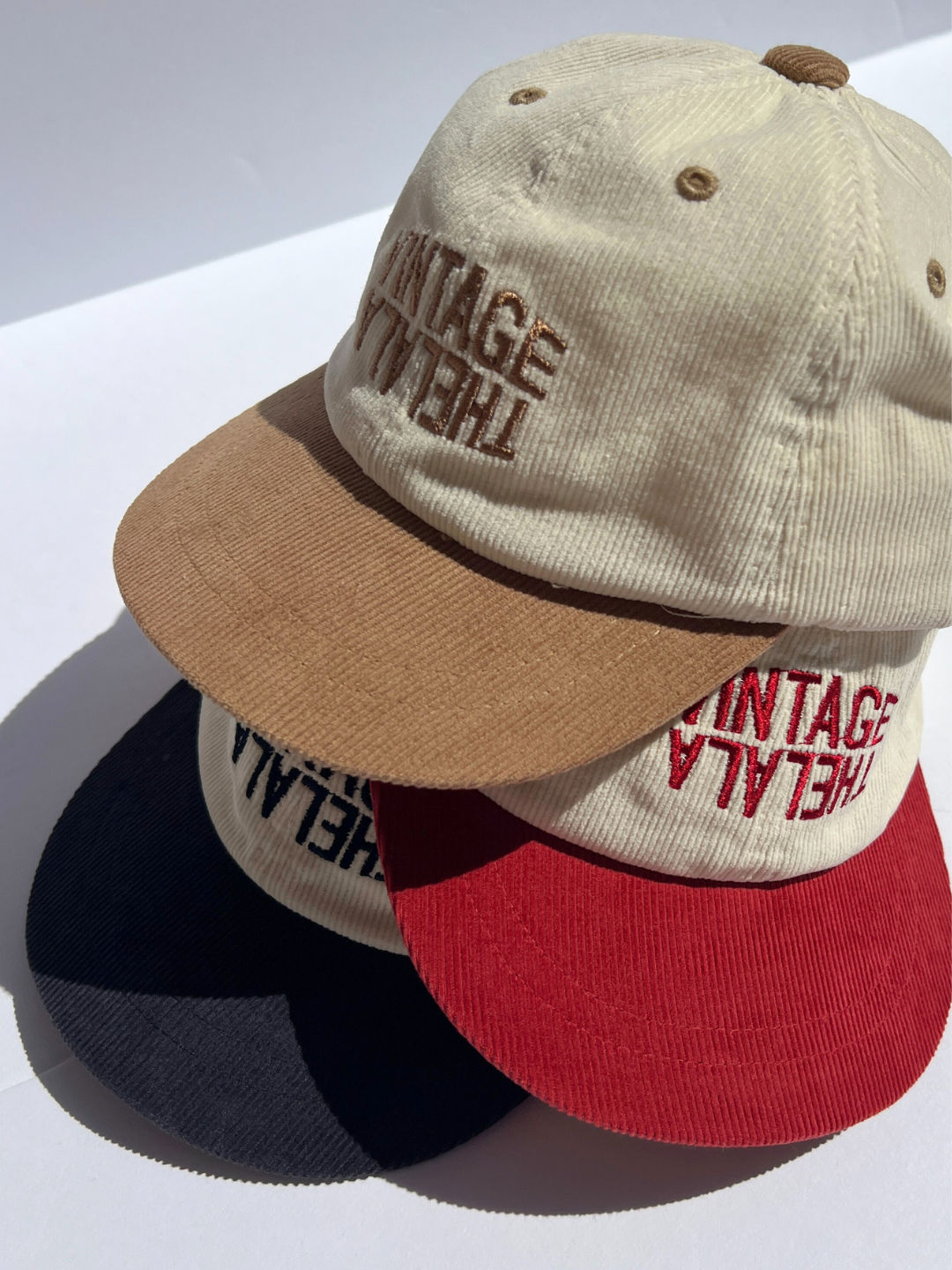 Tan | Three LALA CORDUROY CAPS in beige, brown, and red with "VINTAGE ITALIA" embroidered on the front are stacked on a white surface. Each cap offers an adjustable fit for comfort.