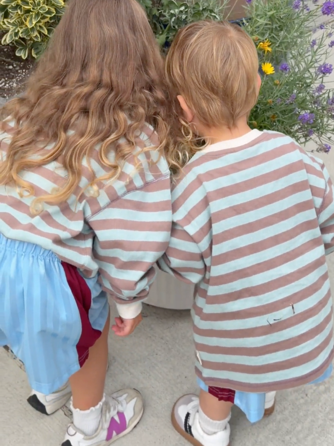Two children seen from behind wearing matching long-sleeve striped shirts with dusty brown and light blue horizontal stripes, standing outdoors near plants.