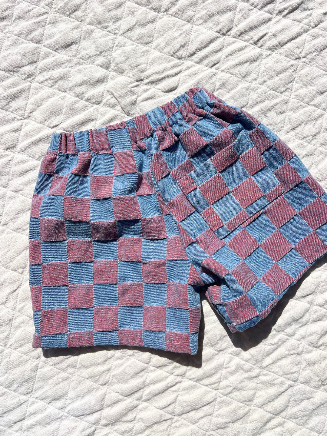 Pink | The TWO TONE DENIM CHECK SHORTS feature a blue and mauve checkerboard jacquard, elastic waistband, and side pocket, shown flat on a quilted white surface. Inspired by a stylish Korean kids brand.