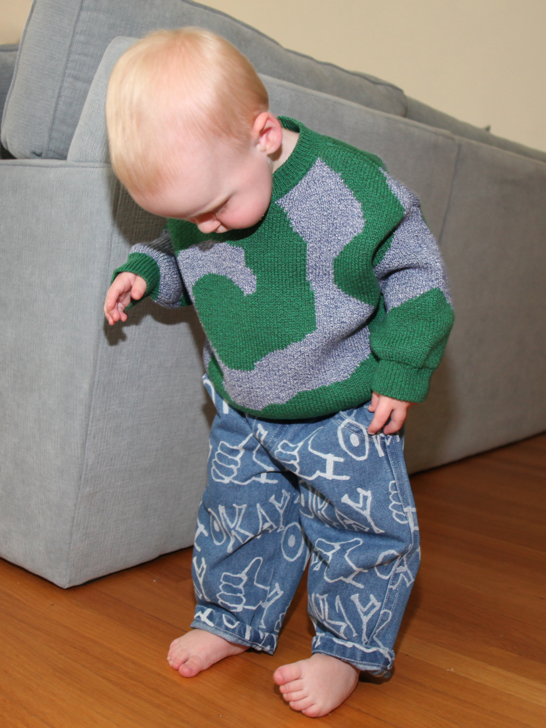 A blonde-haired toddler stands barefoot on a wooden floor in front of a gray sofa, wearing relaxed-fit OKAY JEANS with white graphic designs and a green and gray sweater.