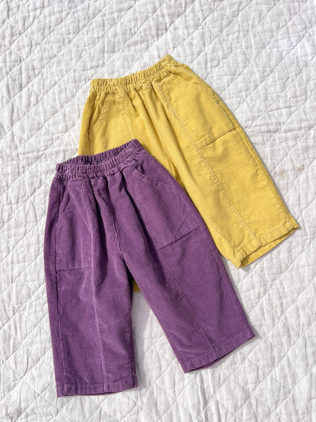 Eggplant | Two corduroy pants, yellow and purple, on a white quilt