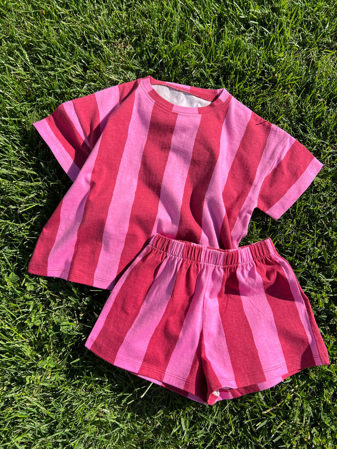 The UMBRELLA STRIPE SHORT SET is a vibrant unisex ensemble featuring a short-sleeved shirt and shorts with bold pink and red stripes in organic cotton—ideal for playful days.