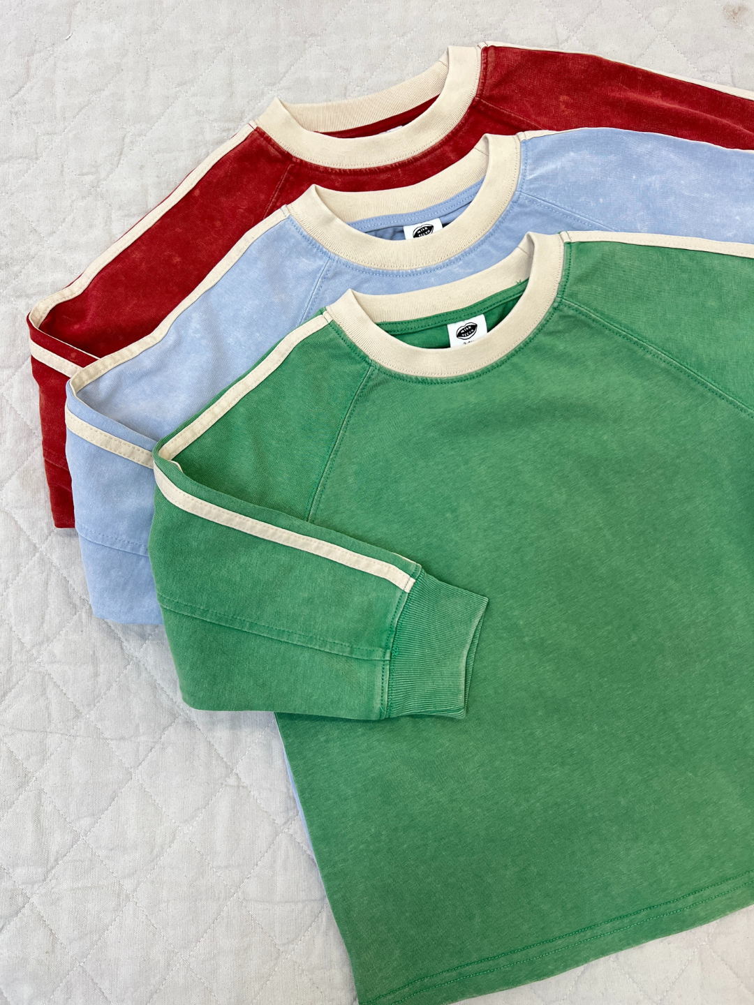 Green | Three HALFTIME LONGSLEEVE tees in green, light blue, and red with cream trim and stripe details are displayed on a quilted white surface, capturing a vintage vibe.