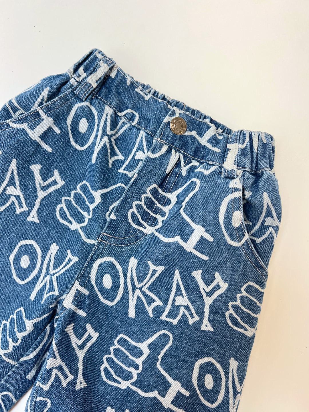 OKAY JEANS: Blue printed denim pants with an elastic waistband, front button, and all-over white "OKAY" text and thumbs-up pattern. These relaxed-fit jeans combine style and comfort.