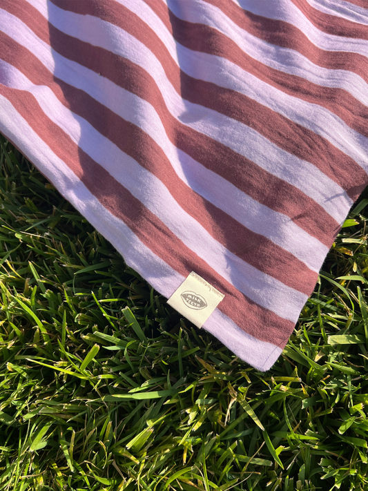 Second image of Purple Stripe | A purple and maroon striped organic cotton swaddle is shown rolled up with a white Milk Teeth label around the middle, on a light grey background.