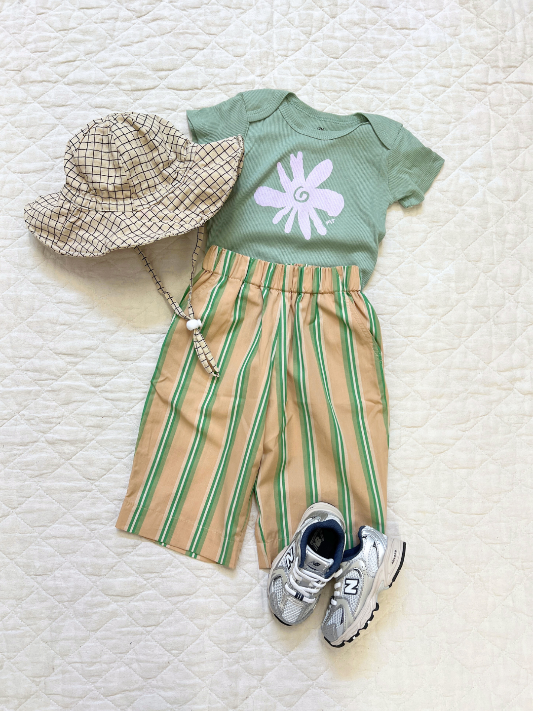 Latte | A flat outfit with the baby off the grid hat in latte, paired with beige and green striped pants, a green flower onesie and grey sneakers, laid on a white cloth.