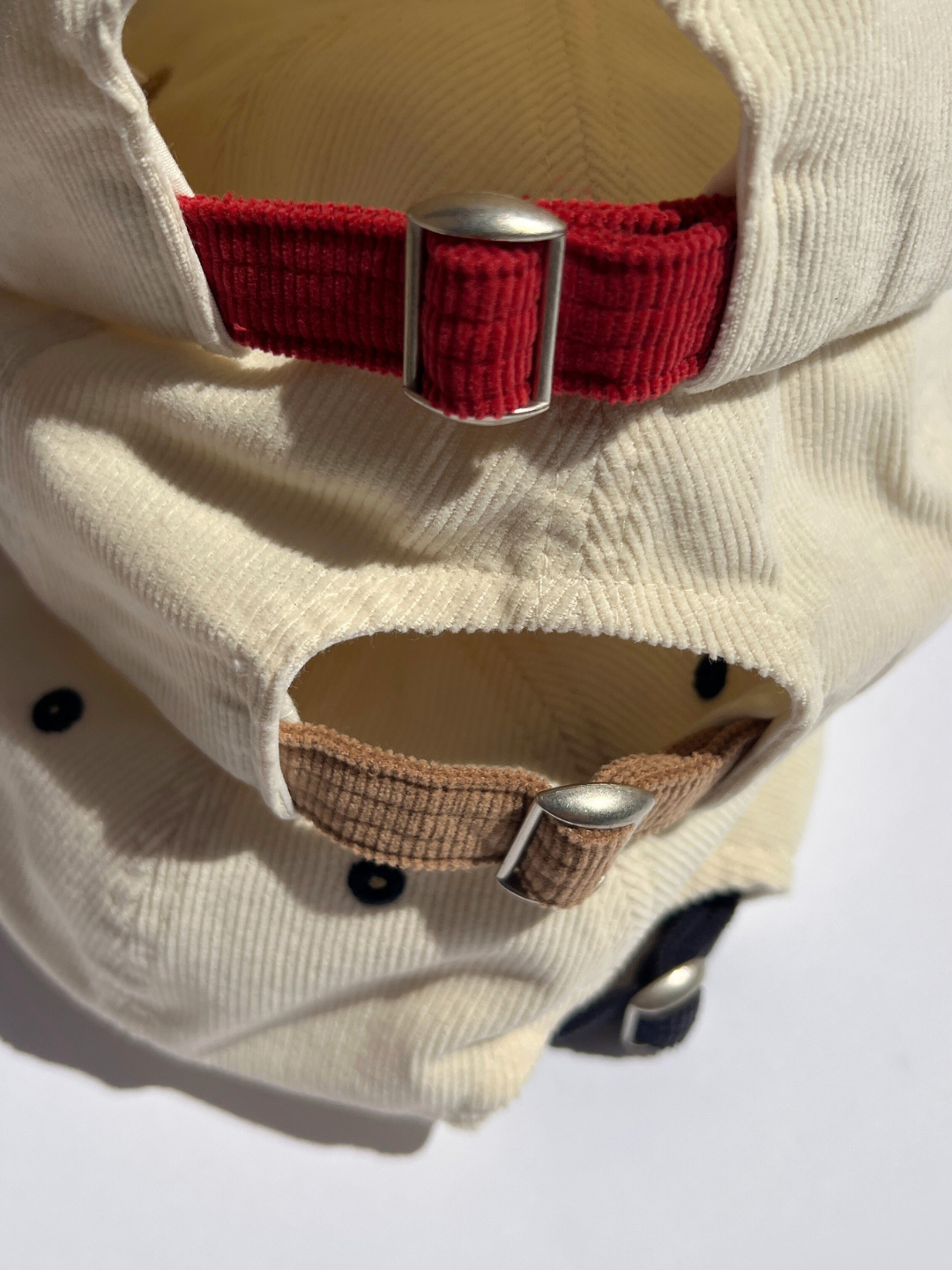 Navy | Close-up of the adjustable fit straps and metal buckles on the back of three LALA CORDUROY CAPS in cream, red, and brown—each toddler baseball cap stacked neatly on top of the other.